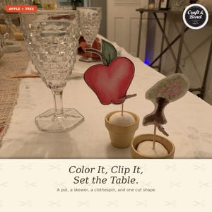 May include: Two small beige pots each containing a white spherical base, a wooden skewer, and a miniature wooden clothespin. One clothespin holds a red apple cutout with a green leaf, and the other holds a cutout of a tree with pink blossoms.