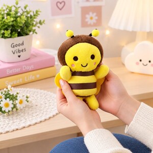 May include: A small plush bee toy featuring a yellow and brown striped body, a brown head with two antennae, black eyes, and a smiling mouth. The soft stuffed animal has small yellow wings on its back and pink circular accents on its cheeks.