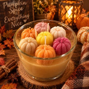 May include: A clear glass bowl filled with wax, containing seven small pumpkin-shaped candles in shades of orange, yellow, pink, and white. Each pumpkin candle features a central white wick. The bowl sits on a circular wooden slice.