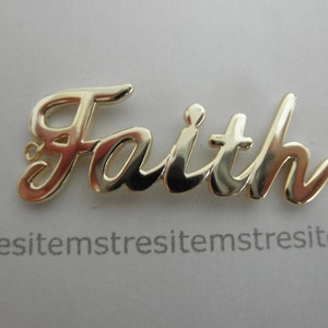May include: Gold-colored metal charm with the word "Faith" written in a cursive script.