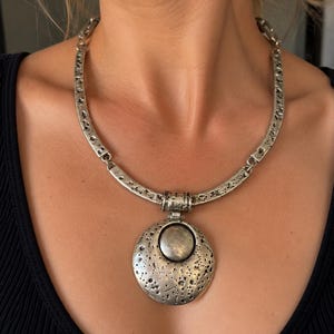 Op de afbeelding: Een zilverkleurige metalen ketting met een stijve, getextureerde halsketting en een grote, ronde hanger. De hanger heeft een kraterachtig, maanachtig oppervlak met een glad, verhoogd midden. Het metaal heeft een donkere, geoxideerde afwerking.