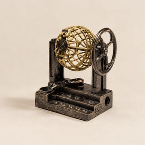 Könnte beinhalten: Eine Lotterie-Ballmaschine aus Metall im Vintage-Stil mit einem dunklen, verwitterten Finish an Basis und Rahmen. Ein goldfarbener Drahtkäfig sitzt zwischen zwei aufrechten Stützen, ergänzt durch ein seitliches Handkurbelrad und eine kleine Ablage.