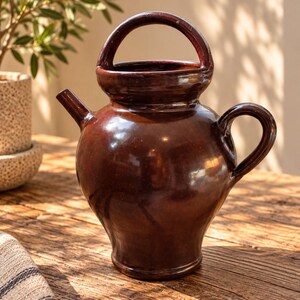 May include: A dark brown glazed ceramic pitcher with a rounded body, a small spout on the left side, a curved handle on the right side, and a large arched handle extending over the top opening. The surface has a smooth, reflective finish.