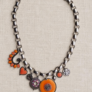 Op de afbeelding: Een ketting van gunmetal-kleurige schakels met diverse bedels, waaronder een oranje paisley-vorm, een oranje hart, een ronde bedel met roze strass-steentjes, een grote oranje schijf met een filigraan centrum en een kleine engel-bedel.