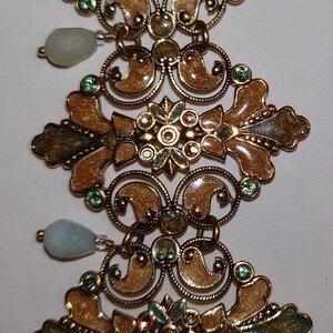 Op de afbeelding: Een verticaal decoratief metalen stuk met vier herhalende, sierlijke goudkleurige filigraansegmenten verbonden door ringetjes. Elk segment bevat een centraal bloemmotief, krulpatronen, kleine groene kristallen en twee lichtblauwe druppelvormige kralen.