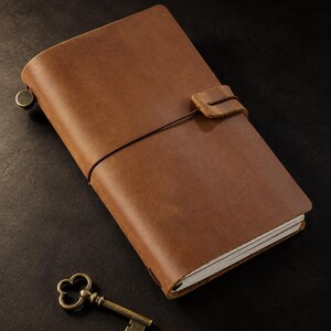 May include: A brown leather journal with a wrap-around elastic cord closure and a small metal fastener on the side, accompanied by a small, ornate brass-colored skeleton key placed near the bottom corner of the notebook.