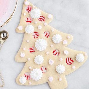 May include: A Christmas tree-shaped sugar cookie decorated with white meringue drops, pink star and rose-shaped sprinkles, and red and white peppermint candies. The cookie is on a white marble surface.