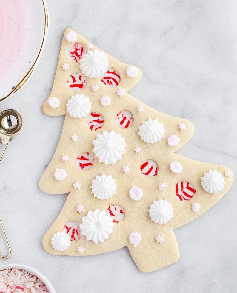 May include: A Christmas tree-shaped sugar cookie decorated with white meringue drops, pink star and rose-shaped sprinkles, and red and white peppermint candies. The cookie is on a white marble surface.