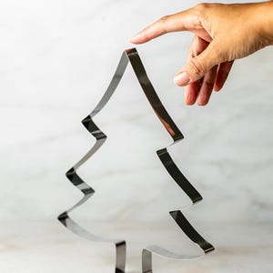 May include: A shiny, silver Christmas tree-shaped cookie cutter. The cutter is made of metal and has a simple, modern design. The tree is held by a hand, with a finger touching the top of the tree.