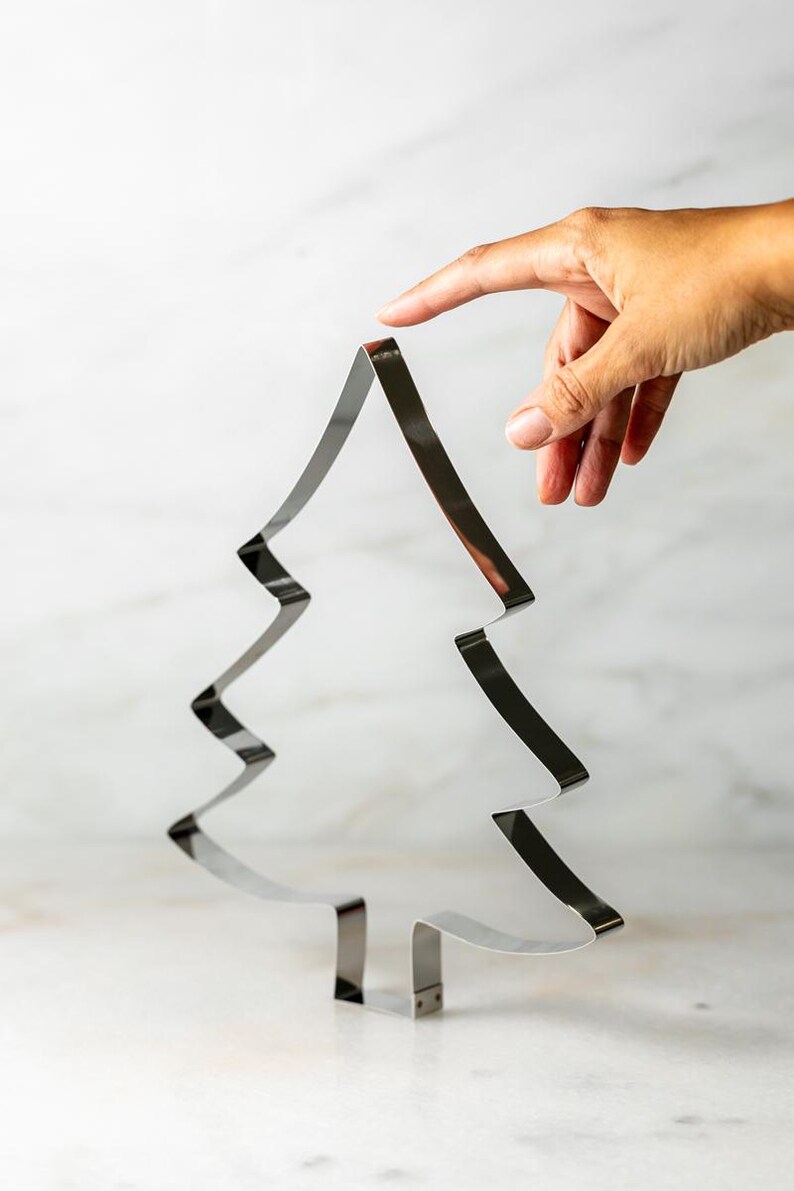 May include: A shiny, silver Christmas tree-shaped cookie cutter. The cutter is made of metal and has a simple, modern design. The tree is held by a hand, with a finger touching the top of the tree.