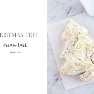 May include: A Christmas tree-shaped cream tart decorated with white frosting, edible flowers, and star-shaped cookies. The tart sits on white parchment paper. Text on the image reads "CHRISTMAS TREE cream tart #cookiecake".