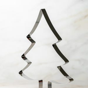 May include: A shiny, silver-colored Christmas tree-shaped cookie cutter. The cutter is made of metal and has a simple, clean design. The tree shape has a pointed top and zig-zag edges. It stands upright on a white surface.