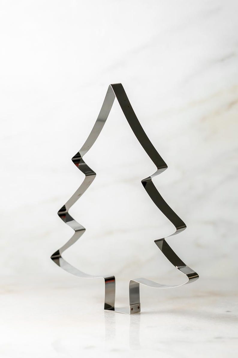 May include: A shiny, silver-colored Christmas tree-shaped cookie cutter. The cutter is made of metal and has a simple, clean design. The tree shape has a pointed top and zig-zag edges. It stands upright on a white surface.