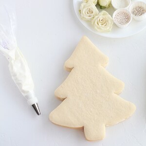 May include: A Christmas tree-shaped cookie sits on a white surface, ready for decorating. A piping bag filled with white frosting and a plate of white roses and various sprinkles are nearby. Baking and decorating supplies.