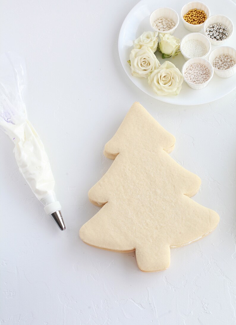May include: A Christmas tree-shaped cookie sits on a white surface, ready for decorating. A piping bag filled with white frosting and a plate of white roses and various sprinkles are nearby. Baking and decorating supplies.