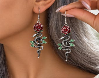 May include: A pair of silver-toned drop earrings featuring a detailed snake coiled around a stem with green leaves and a vibrant red rose at the top. The earrings hang from silver hooks and showcase intricate metalwork with enamel color accents.