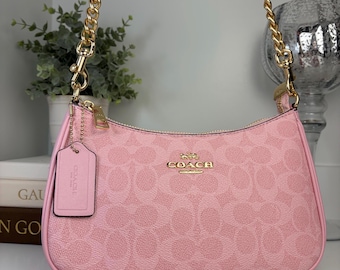 Könnte beinhalten: Eine hellrosa Coach-Handtasche mit dem charakteristischen Monogramm-Muster in einem helleren Rosaton. Die Tasche verfügt über einen goldfarbenen Kettenriemen, einen passenden rosa Lederanhänger und goldfarbene Beschläge, inklusive Coach-Logo.