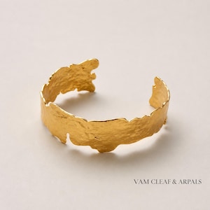 Può includere: Un bracciale a fascia in oro con una superficie testurizzata e martellata dai bordi irregolari e organici. Il metallo ha una finitura brillante e riflettente. Il testo VAM CLEAF & ARPALS è stampato nell'angolo in basso a destra.