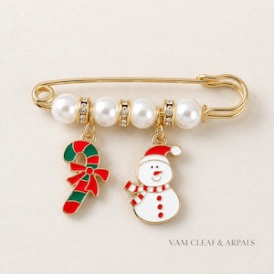 May include: A gold-tone safety pin brooch featuring four white faux pearls separated by small crystal-encrusted rings. Two charms hang from the pin: a red and green candy cane with a bow and a white snowman with a red hat and scarf. Text reads VAM CLEAF & ARPALS.
