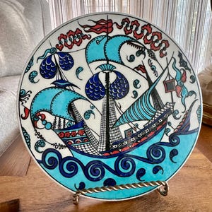 May include: A round ceramic decorative plate featuring a hand-painted illustration of a sailing ship on turquoise water. The design includes intricate black line work, blue and red accents, and stylized cloud motifs against a white background.