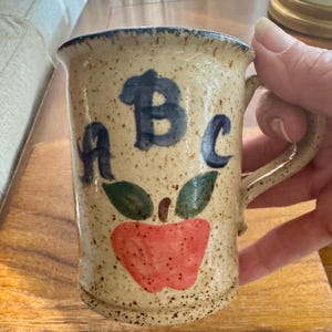 May include: A speckled beige ceramic mug featuring the hand-painted blue letters A, B, and C arranged above a red apple illustration with two green leaves. The mug has a sturdy handle and a slightly flared rim with a dark speckled edge.