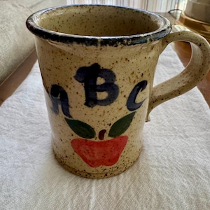 May include: A speckled beige ceramic mug with a dark blue rim and handle. The front features the hand-painted blue letters a, B, and c above a red apple illustration with two green leaves. The mug has a cylindrical shape and a sturdy side handle.