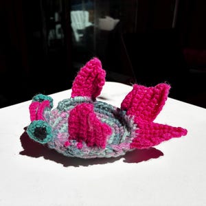 May include: A crocheted hammerhead shark toy featuring a variegated light blue and grey body with vibrant hot pink fins and tail. The shark has two prominent eyes on the sides of its wide, flat head, crafted with black yarn centers.