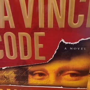 Könnte beinhalten: Das Buchcover von The Da Vinci Code von Dan Brown zeigt einen tiefroten Hintergrund mit großen, geprägten goldenen Buchstaben. Ein zerrissener Bereich enthüllt eine Nahaufnahme menschlicher Augen und handschriftlicher Notizen, mit dem Text Author of Angels & Demons.