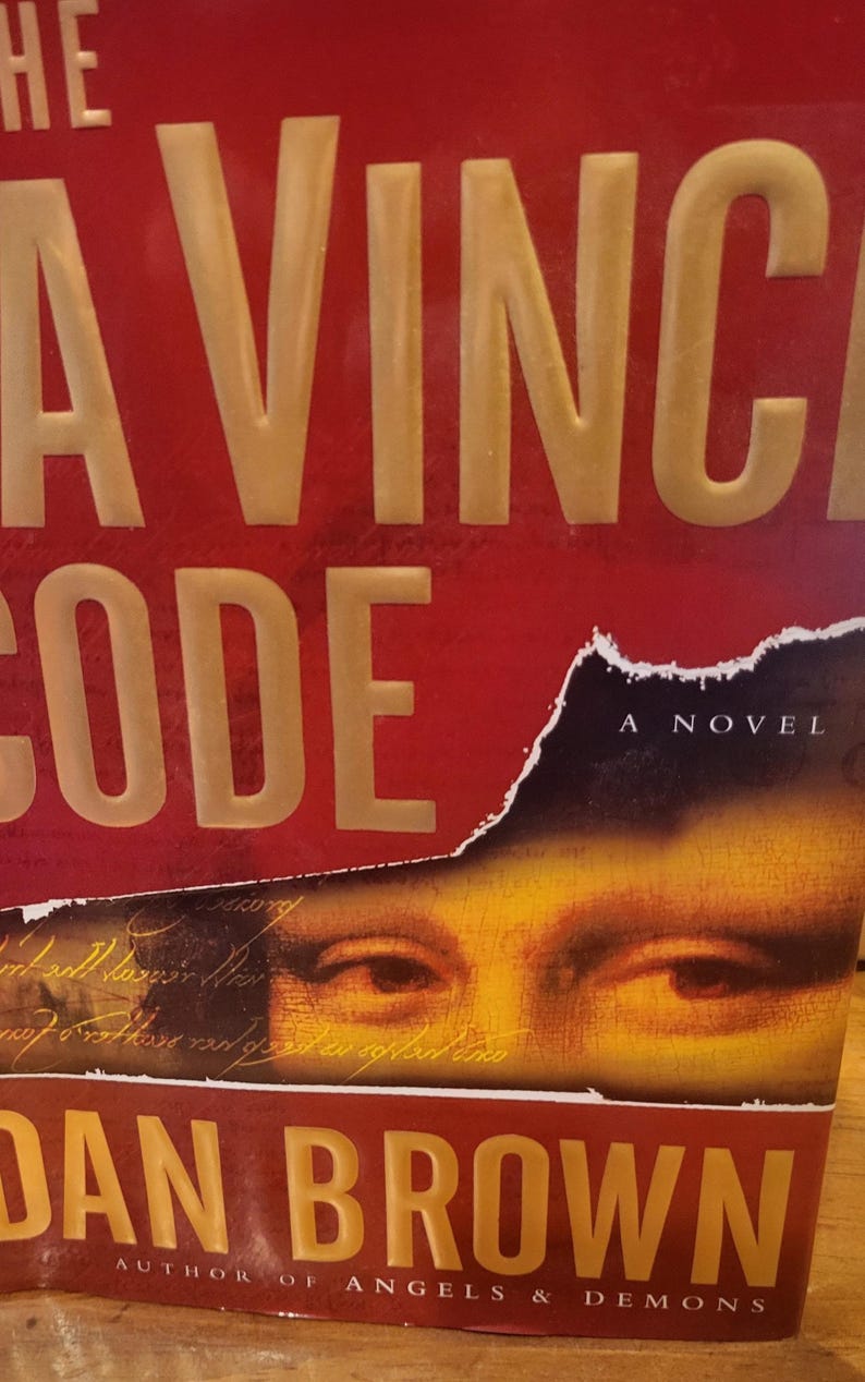 May include: The book cover for The Da Vinci Code by Dan Brown features a deep red background with large, embossed gold lettering. A torn section reveals a close-up of human eyes and handwritten script, with the text Author of Angels & Demons below.
