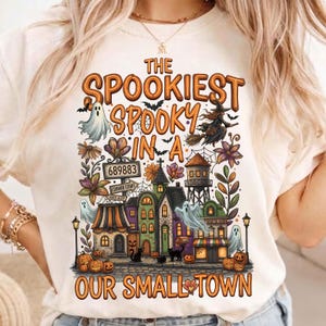 Puede incluir: Una camiseta color crema con un gráfico colorido de Halloween y el texto THE SPOOKIEST SPOOKY IN A OUR SMALL TOWN. El diseño incluye fantasmas, una bruja en una escoba, una torre de agua, calabazas, gatos negros y señales de calle.