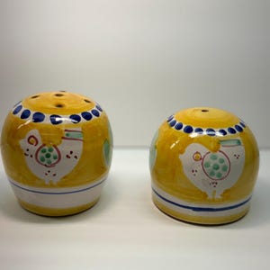 May include: Two rounded ceramic shakers featuring a yellow base with hand-painted white chicken motifs, green polka-dot accents, and a blue dotted border around the top. Each shaker has a white bottom section with a thin blue horizontal stripe.