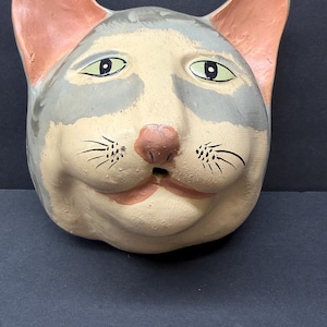 May include: A ceramic cat head wall plaque featuring a cream-colored face with grey patches around the eyes and ears. The cat has large almond-shaped green eyes, a pink nose, and painted black whiskers. The ears are painted a soft pink color.