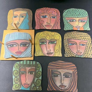 May include: A set of seven flat, square-shaped coasters featuring stylized illustrations of faces with diverse hair colors and patterns. One coaster is held in a light-colored wooden stand with four pegs, while the others are arranged around it.