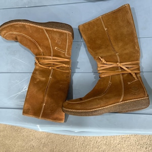 May include: A pair of brown suede boots with laces that wrap around the ankles. The boots have a rounded toe and a low heel.