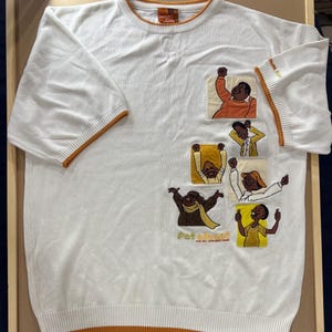 May include: White short-sleeve shirt with orange trim and patches featuring cartoon characters. The patches depict various figures in different poses. The text "Fat Albert and the Junkyard Gang" is visible on the shirt.