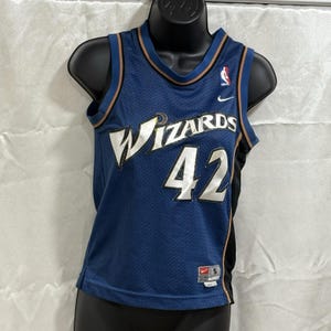 Puede incluir: Una camiseta de baloncesto azul de los Washington Wizards con la palabra "Wizards" en escritura blanca en el pecho y el número "42" en blanco. La camiseta tiene un ribete marrón y dorado y el logo de Nike.