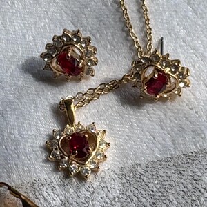 May include: A gold-toned jewelry set featuring a necklace, earrings, and a pendant. Each piece has a central red gemstone surrounded by smaller clear stones. The necklace and earrings have a floral design.