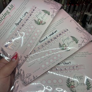 May include: A press-on nail sizing kit featuring three clear plastic cards labeled Almond-Long, Almond-Medium, and Almond-Short. Each card displays numbered nail tips from 0 to 9, circular spaces for finger measurements, and a frog fern logo.
