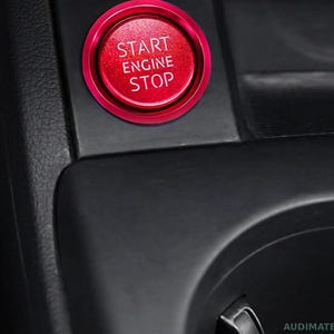 May include: A circular red engine start stop button with a metallic finish, featuring the white text START ENGINE STOP printed in the centre. The button is mounted within a black plastic console panel inside a vehicle interior.