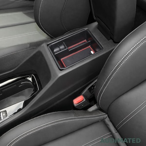 May include: A black plastic car centre console organiser tray featuring red trim accents and a textured non-slip base. The insert is positioned within the vehicle's centre storage compartment between the front seats, which have black upholstery.