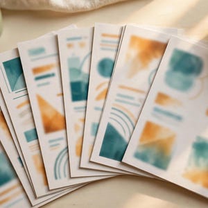May include: A collection of white rectangular paper sheets arranged in a fan shape, each featuring abstract watercolor designs in shades of teal and mustard yellow, including geometric shapes, lines, and soft blended color patches.