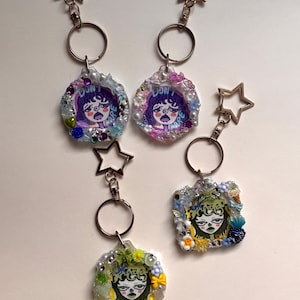 May include: Three keychains featuring star-shaped clasps and circular or square resin charms. The charms are decorated with small beads, flowers, and illustrations of faces with the text "DON'T CARE" or "OKAY" in purple, green, and yellow tones.