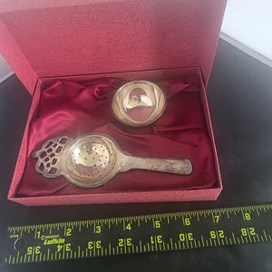 May include: A silver-toned tea strainer and a small matching bowl presented in a red textured box with a satin lining. A black Lufkin ruler is placed in front, showing the strainer measures approximately 6 inches in total length.