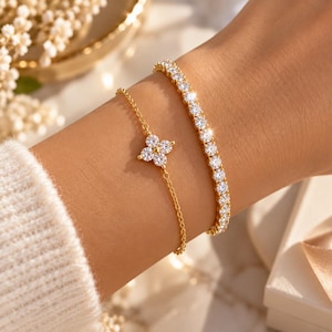 May include: A set of two gold-toned bracelets. One is a delicate chain featuring a four-petaled flower charm set with clear stones. The second is a tennis-style bracelet featuring a continuous row of clear stones set in a gold-toned metal band.