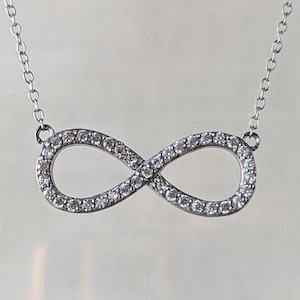 May include: A silver-toned necklace featuring an infinity symbol pendant encrusted with small, clear, sparkling stones. The pendant is attached to a delicate, thin silver-toned chain that extends upwards from both sides of the infinity loop design.