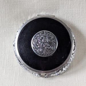 May include: A round compact mirror featuring a black center inlay surrounded by a silver-toned metal frame. The center of the black surface is adorned with a circular silver-toned medallion embossed with a detailed floral pattern of roses.