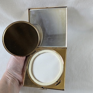 May include: A square gold-toned metal compact case featuring a hinged lid with an attached circular mirror. The interior contains a white circular powder puff nestled within a square frame. The metal finish is polished and reflective throughout the piece.