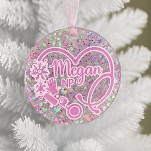 May include: A round, pink and glittery ornament with the name "Megan NP" in a heart-shaped design. The ornament includes a stethoscope, snowflake, and medical gloves. A pink ribbon is attached for hanging.