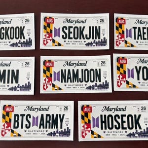 以下が含まれることがあります： AUG、26、Maryland、Baltimoreの文字と、JUNGKOOK、SEOKJIN、TAEHYUNG、JIMIN、NAMJOON、YOONGI、BTS ARMY、HOSEOKの名前が記載された7枚の長方形のメリーランド州ナンバープレートステッカー。各ステッカーには紫色のロゴと都市のスカイラインが描かれています。