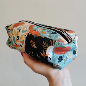 May include: A rectangular fabric zip pouch featuring a patchwork design with various autumn-themed patterns. The fabric displays orange pumpkins, brown oak leaves, and white berries on backgrounds of light blue, cream, and black. A silver metal zip.